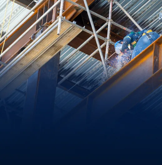 Worker in blue protective gear welding steel beams on scaffolding with sparks flying