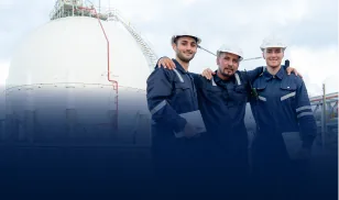 Three workers in navy overalls and hard hats smiling in front of a large gas storage tank